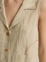 Camisa sin mangas de lino, color blanco, con cuello solapa, cierre frontal con botones y bolsillo pequeño en el pecho. Presenta costuras decorativas a la vista que definen la silueta.