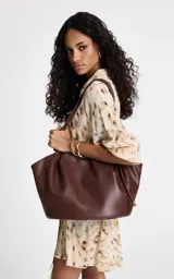 Cartera tipo tote de gran tamaño color chocolate, con diseño flexible y cierre de cordón ajustable. Presenta un acabado liso y minimalista con el logo de la marca grabado en la parte frontal.