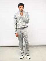 Conjunto deportivo Umbro compuesto por campera de cuello alto con cierre frontal y pantalón de corte recto. La campera presenta bloques de color blanco en los hombros y el logo de la marca bordado en el pecho. Confeccionado en tejido sintético con acabado satinado.