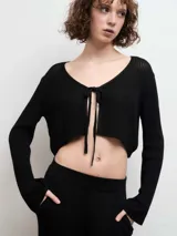 Cardigan de punto negro, de corte corto y diseño abierto con cierre de lazo frontal. Presenta mangas largas y una textura acanalada fina.