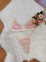 Conjunto de lencería compuesto por un bralette triangular y una tanga, confeccionados en algodón color rosado con detalles de encaje en tono crema. El bralette cuenta con breteles ajustables y el conjunto presenta terminaciones delicadas.