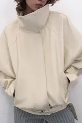 Chaqueta bomber oversize de efecto piel en color crema, con cuello subido, manga larga con hombro caído y cintura con frunces y cinturón ajustable con hebilla metálica. Cuenta con cierre frontal oculto y bolsillos laterales de vivo.