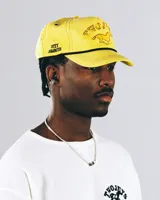 Gorra tipo trucker color amarillo con bordado frontal que incluye la frase 'TWOJEYS' y la silueta de un caballo. Presenta un cordón decorativo negro en la base de la visera y el texto 'CITY COWBOYS' bordado en el lateral.
