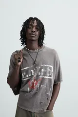 Camiseta gris de punto con efecto lavado y roturas, corte "boxy fit", cuello redondo y manga corta. Presenta un estampado frontal en blanco y rojo con diseño abstracto.