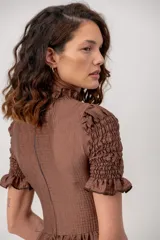 Vestido corto color marrón chocolate, con cuerpo fruncido tipo nido de abejas, cuello alto con volados y mangas cortas abuchonadas con terminación en volado. Presenta falda evasé con vuelo.