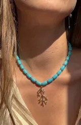 Collar corto de cuentas cilíndricas de piedra turquesa intercaladas con pequeños discos de acero, con un dije colgante plateado en forma de rama o coral.