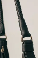 Cartera tipo hobo de gamuza color marrón, con correa trenzada de cuero negro y detalles laterales en cuero negro.