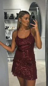 Vestido corto de fiesta color bordó, confeccionado en tela con lentejuelas. Presenta breteles finos, escote redondo y un diseño con volado en el ruedo.