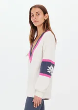 Sweater de punto color crema, con escote en pico con ribete rosa y gris. Presenta mangas largas con franjas grises y rosas en los puños, y logo bordado en el brazo.