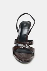 Sandalia de tacón alto fino, color marrón oscuro con textura de relieve de piel de serpiente. Presenta tiras anchas cruzadas en la parte delantera y una tira trasera con elástico ajustable. La punta es cuadrada.