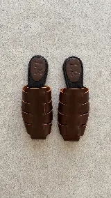 Zuecos de cuero marrón con diseño de tiras anchas entrelazadas estilo romano, punta cuadrada y plantilla plana negra con logo grabado de la marca.