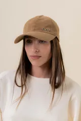 Gorra de diseño clásico con visera curva, confeccionada en material símil gamuza color tostado. Presenta un logo metálico con la palabra 'peach' en el frente y cuenta con un sistema de ajuste posterior.