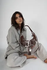 Bolso marrón con efecto desgastado y detalles pintados en blanco. Tiene forma de media luna, correa de hombro y cierre con hebilla plateada.