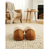 Pantuflas de gamuza color beige con plataforma y forro interior de corderito sintético. Presentan un diseño sin talón con el nombre de la marca bordado en el lateral.