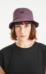 Piluso de estilo bucket hat en color violeta oscuro, con costuras circulares en el ala y parche cuadrado frontal con logo de tijeras.