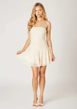 Vestido corto strapless con acabado de lentejuelas brillantes en color crema. Presenta un diseño ajustado en el torso con detalle de frunces laterales y una falda acampanada con vuelo.