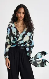 Blusa de tejido fluido con estampado abstracto en tonos azules, negros y beige. Presenta un diseño cruzado con escote en V, mangas 3/4 anchas y un lazo lateral ajustable que define la silueta.