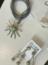 Collar artesanal de la marca Fluss con dije en forma de sol hecho de bronce y alpaca, con incrustaciones de piedra en tono verde y centro de resina con fragmentos. Viene con una tira de gamuza gris.