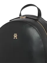Mochila negra de poliuretano con forma redondeada en la parte superior y base rectangular. Cuenta con asa de mano superior, correas acolchadas ajustables y un logotipo metálico monograma TH en el centro.