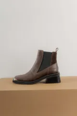 Botas tipo chelsea de cuero color marrón oscuro con acabado brillante. Presentan punta cuadrada con detalle de puntera metálica, elásticos laterales y taco cuadrado bajo.
