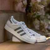 Championes Adidas Superstar de diseño clásico, color blanco con las icónicas tres tiras laterales en tono gris oscuro. Presenta detalle de estampado animal print en el talón y puntera de goma reforzada.