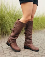 Botas de caña alta confeccionadas en cuero color marrón caoba. Presentan un diseño robusto con tres hebillas metálicas ajustables en el lateral superior y una correa con hebilla y remaches metálicos en la zona del tobillo. Cuentan con un taco bajo cuadrado y suela de goma resistente.