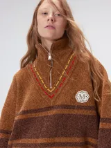 Pullover de punto con textura aterciopelada, diseño oversize, cuello polo con cierre frontal parcial y detalles de pespuntes en contraste. Presenta rayas horizontales en tonos tierra y un parche bordado con la letra M en el pecho.
