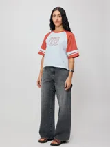 Remera bicolor de ajuste holgado, con cuerpo blanco y mangas raglán en tono terracota con detalles de líneas blancas en los puños. Presenta estampado frontal con el texto "RUSTY 85".