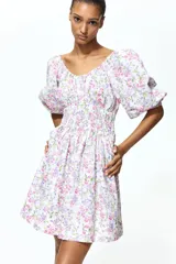 Vestido corto de algodón con estampado floreado en tonos rosados y lilas sobre base blanca. Presenta escote en V fruncido, mangas globo a los codos con puños elásticos y sección fruncida en la cintura.