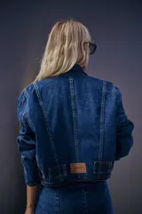 Campera de jean de corte vintage en color azul, con pespuntes en hilo color maíz y detalles de denim negro en los puños, la cintura y los costados. Cuenta con una etiqueta de cuero sintético con el logo de la marca en la parte trasera inferior.