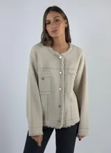 Chaqueta color crudo con cuello redondo, cierre frontal con botones plateados, bolsillos frontales y detalles de flecos en cuello, cierre, bolsillos y ruedo.