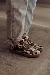 Zapatillas de cuero con estampado que simula pelo de vaca o manchas marrones y blancas. Tienen detalles en cuero marrón liso, cordones marrones y suela de goma color caramelo.