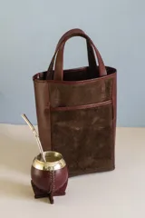 Bolso tipo matera de diseño vertical, confeccionado en una combinación de cuero liso color bordó y gamuza color marrón chocolate. Cuenta con doble asa superior y un bolsillo frontal abierto.
