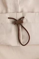 Campera rompevientos de color beige, modelo oversized con cuello alto y capucha con cordón ajustable. Cuenta con cierre frontal marrón, mangas raglán con puños elásticos, bolsillos laterales y detalles de ribetes en tono marrón oscuro.