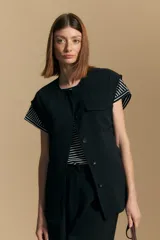 Chaleco negro de corte estructurado con cuello redondo, cierre frontal oculto y lazo ajustable en la cintura. Presenta dos bolsillos frontales con solapa y diseño de hombros caídos.