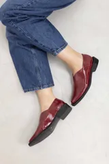 Mocasines de charol color bordó, de diseño vanguardista con corte asimétrico en el empeine, abertura en V, punta fina y suela plana con pequeño tacón.