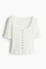 Top de manga corta de punto pointelle suave, color blanco, con ajuste entallado, escote en U y hilera de botones en la parte delantera. Presenta ribete levemente festoneado en el escote y la tapeta.