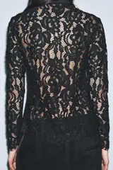 Camisa de manga larga confeccionada en encaje negro con patrón floral. Presenta cuello solapa de tejido liso negro y cierre frontal de botones. El bajo del ruedo y los puños tienen terminación de encaje festoneado.