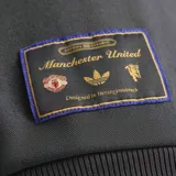 Campera deportiva Adidas del Manchester United, color gris con detalles en dorado. Presenta cierre frontal, cuello alto, bolsillos laterales y escudo del club bordado en el pecho.