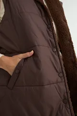 Chaleco acolchado color chocolate con interior y cuello de corderito sintético. Presenta cierre frontal con botones a presión y bolsillos laterales.