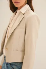 Saco corto tipo blazer en color beige, confeccionado en lino con acabado brillante. Presenta solapas clásicas, cierre frontal con un botón y mangas largas con puños abotonados.
