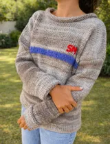 Sweater de punto en tonos beige con efecto degradado, cuello redondo y manga larga. Presenta una franja horizontal en color azul y el bordado de las letras 'SM' en color rojo en el pecho.