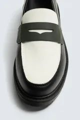 Mocasín de cuero marrón oscuro con antifaz en tono camel. Presenta horma redonda y suela track gruesa de goma negra.