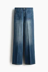 Pantalón de denim azul con lavado medio, corte de pierna recta y tiro alto. Presenta bolsillos de parche delanteros con solapa, trabillas para cinto y detalles de pliegues marcados en el centro de las piernas.