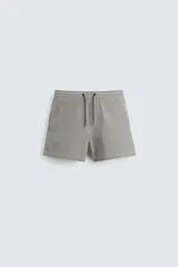 Bañador de hombre color beige con finas rayas verticales oscuras, confeccionado en tejido técnico con textura seersucker. Presenta cintura elástica ajustable con cordón negro, bolsillos laterales y un bolsillo trasero de vivo.