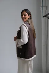 Chaleco de cuero sintético color chocolate con interior y bordes de corderito beige. Presenta solapas amplias, dos bolsillos frontales con detalle de corderito y corte recto.