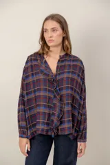 Camisa de corte holgado con estampado de cuadros escoceses en tonos azul, rojo y marrón. Presenta cuello mao, cierre frontal con botones y un detalle de volado vertical a lo largo de la cartera. Mangas largas con puños abotonados.