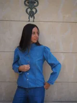 Camisa de manga larga confeccionada en broderie blanco, con diseño entallado, cuello alto y cierre frontal de botones. Presenta mangas abullonadas con hombros fruncidos y puños terminados en volados.