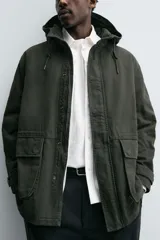 Parka color verde militar de corte relaxed fit, confeccionada en tejido de algodón con interior acolchado. Cuenta con cuello subido, capucha ajustable con cordón, cierre frontal de cremallera oculta por solapa con botones a presión y amplios bolsillos frontales de plastrón con solapa.