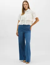 Camisa de algodón blanca con cuello clásico y cierre frontal de botones. Presenta un diseño con capas superpuestas y volados, combinando paneles de tela lisa con tejido bordado suizo de motivos calados y una cenefa decorativa. Posee mangas cortas y una silueta acampanada.